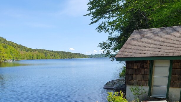 A lakeside cabin with a wooden exterior and a green door, surrounded by clear water and lush vegetation. - Stunning Belgrade Lakes Waterfront Retreat – Dock, Kayaks & Breathtaking Views! (Rome)