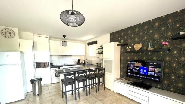 A modern kitchen with white cabinets, a dining area with four chairs, a black pendant light, and a TV on a white stand. - Les Calanques, one-bedroom apartment with air conditioning, large terrace and elevator (Cavalaire-sur-Mer)