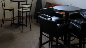 Un petit espace repas, à l’abri, avec une table ronde et des chaises, un grand lampadaire et une fenêtre donnant sur un paysage verdoyant.