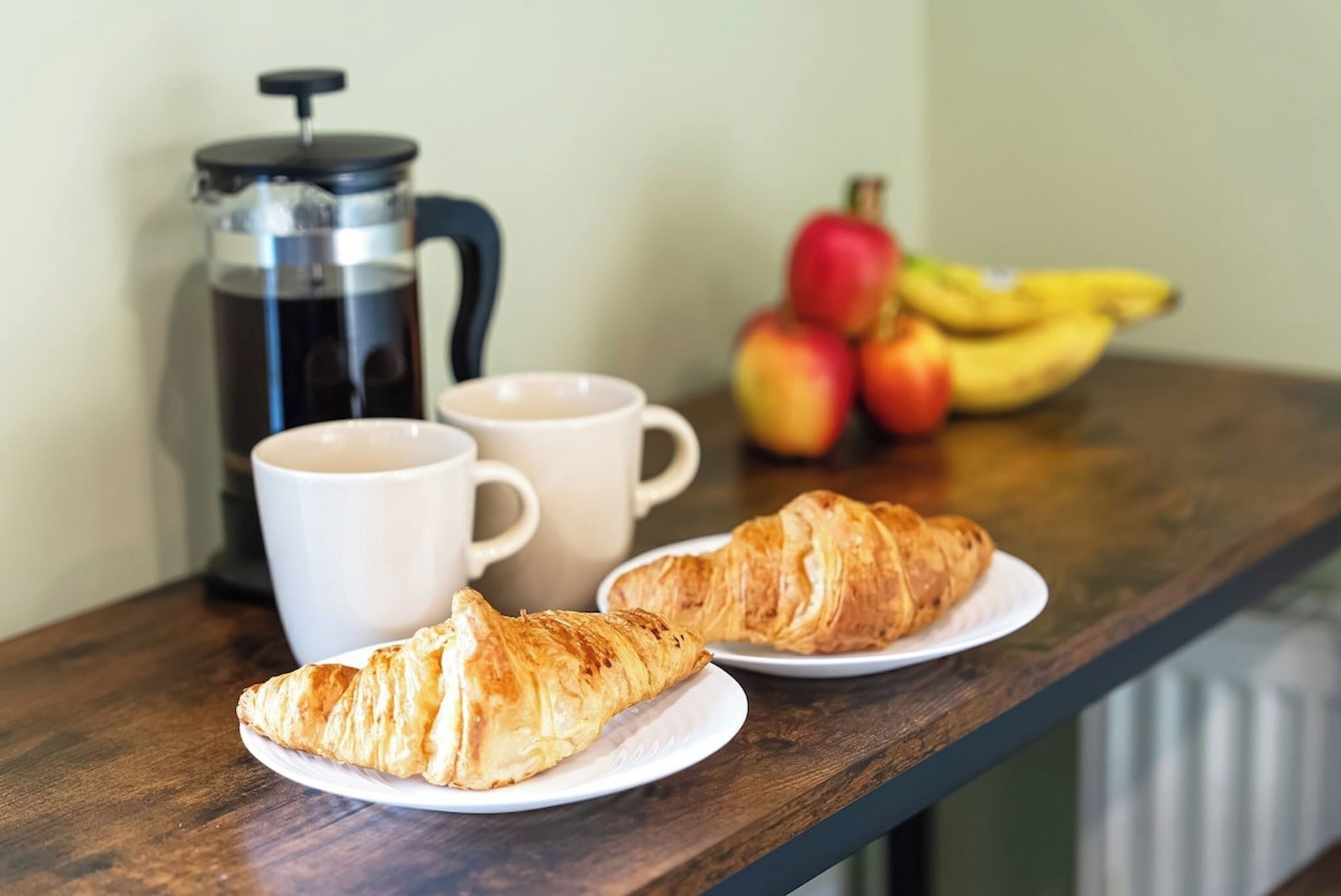 Un tavolo di legno con due croissant, una caffettiera, due tazze bianche e un'elegante composizione di frutta.