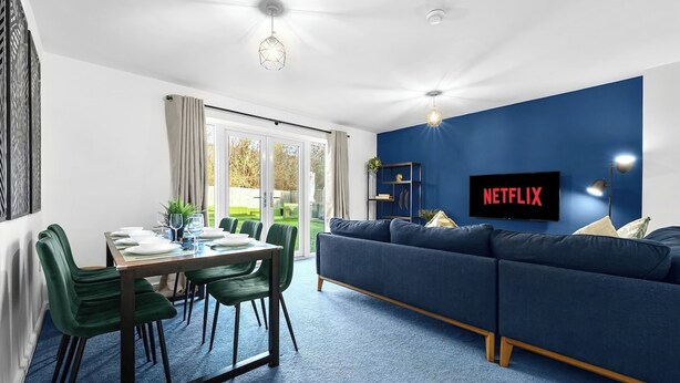 A modern living room with a blue sofa, a dining table with chairs, a TV displaying 'NETFLIX,' and a large window with curtains.