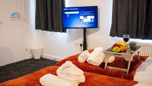A hotel room with a bed, a TV on a stand, a tray with fruits and a glass of wine, and towels on a fluffy orange rug. - The Pines Villa 1 | Spacious 3BR | Cinema, Games & EV | Parking | Sleeps 10+ (West Northamptonshire)