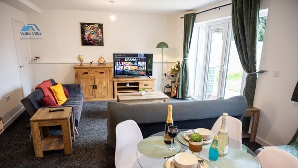 A modern living room with a sofa, a TV, a small dining table with plates and drinks, and a wooden cabinet. - The Pines Villa 1 | Spacious 3BR | Cinema, Games & EV | Parking | Sleeps 10+ (West Northamptonshire)