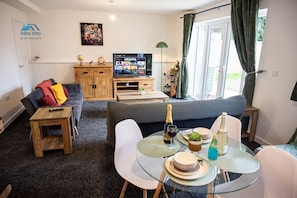 Una sala de estar moderna con un sofá, un televisor, una pequeña mesa de comedor con platos y bebidas, y un mueble de madera.