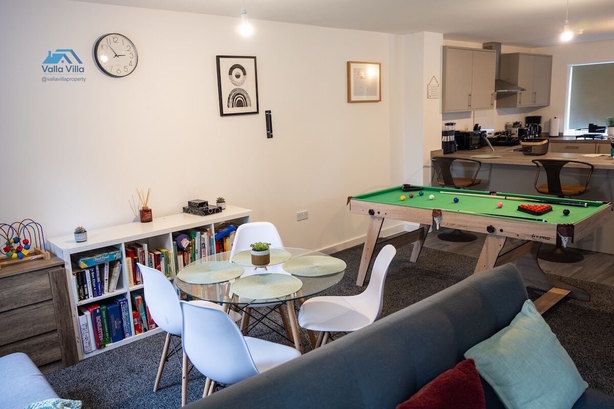 A living room with a pool table, a sofa, a dining table with chairs, a bookshelf, and a kitchen area in the background.