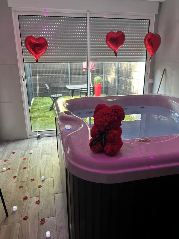Tinaco con un osito de peluche y rosas rojas, rodeado de globos con forma de corazón, con un área de patio visible a través de una puerta corrediza.