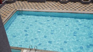 Una piscina rectangular con agua azul cristalina, rodeada por un suelo de baldosas a cuadros y una cerca de metal.