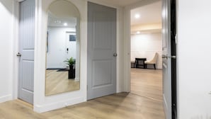 Un couloir avec deux portes blanches, dont l’une est dotée d’un miroir. On aperçoit une plante dans un coin et une chaise au fond.