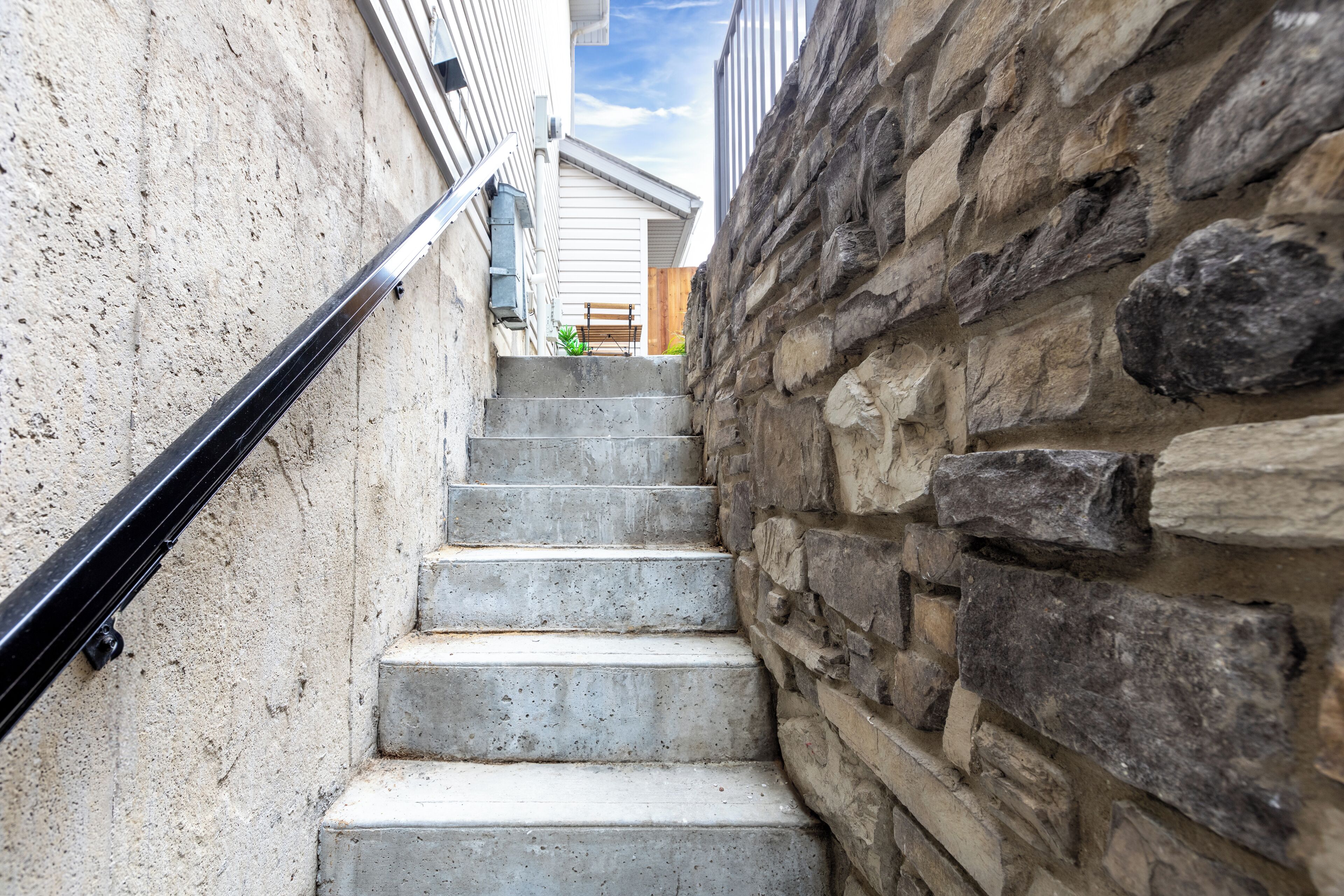 Uma escadaria estreita com corrimão de metal, com uma parede de concreto de um lado e uma parede de pedra do outro.