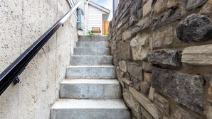 Un escalier étroit doté d’une rampe métallique, bordé d’un côté par un mur en béton et de l’autre par un mur en pierre.