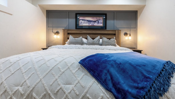 Un lit bien fait, avec une couette blanche matelassée, un plaid bleu et un tableau encadré au-dessus.