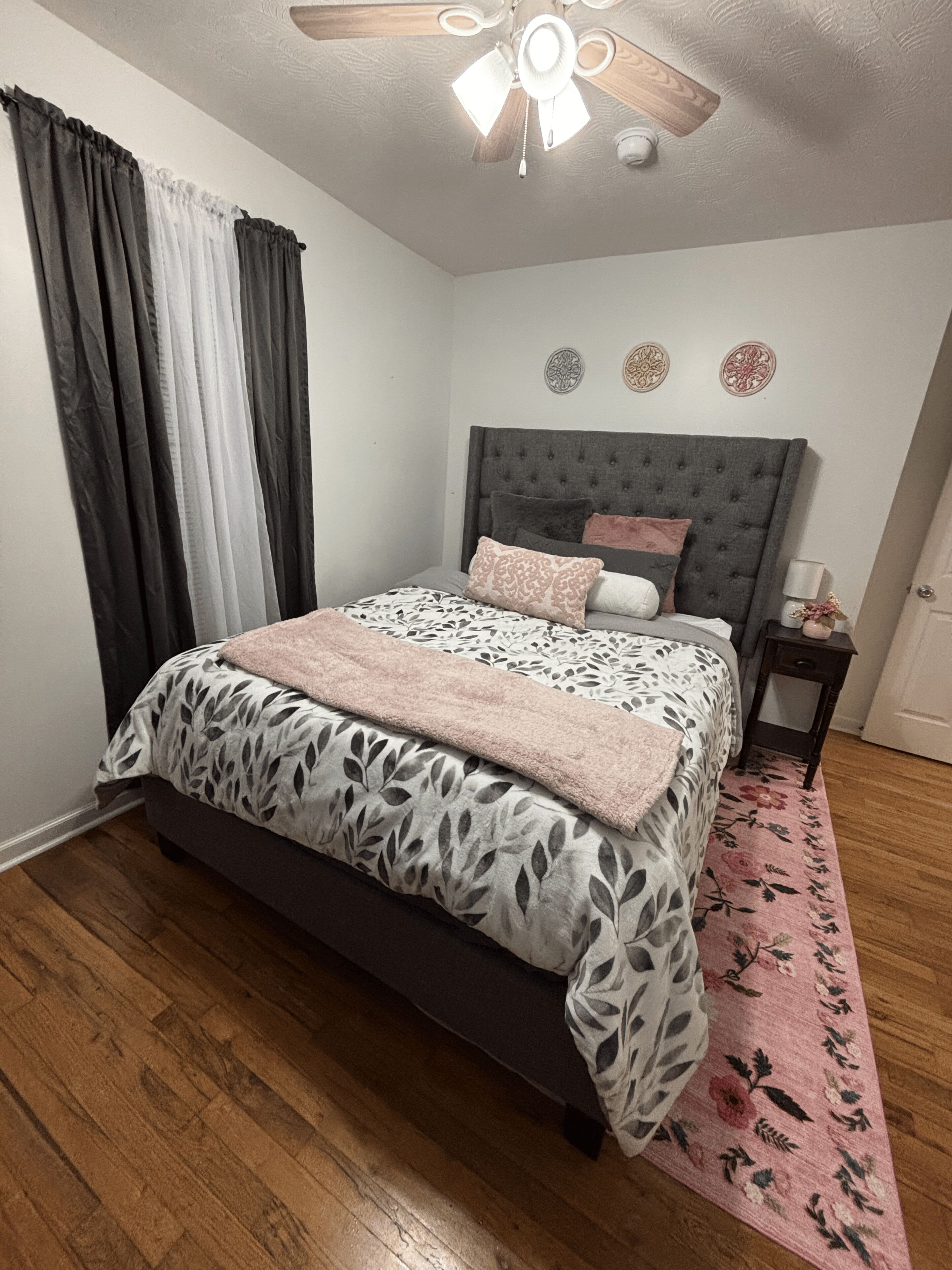 Een slaapkamer met een bed, een plafondventilator, een nachtkastje en een vloerkleed met bloemenmotief.