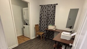 Ett kompakt kök med trägolv, en takfläkt, ett kylskåp, en stol, ett bord med en lampa och mönstrade gardiner.