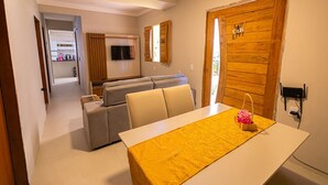 A modern living room with a sofa set, a dining table with a yellow tablecloth, and a wooden door with a coat hanging on it. - Casa com Piscina e Churrasqueira em Arembepe (Camaçari)