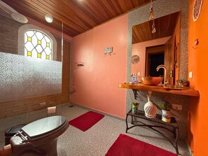 Une salle de bain avec un plafond en bois, un lavabo avec un miroir, une fenêtre à motifs losangés et un tapis rouge.