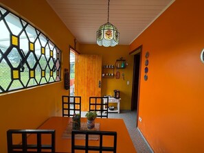 Un espace repas comprenant une table, des chaises, un luminaire suspendu et une vue sur l’extérieur à travers une fenêtre ornée d’un motif géométrique.