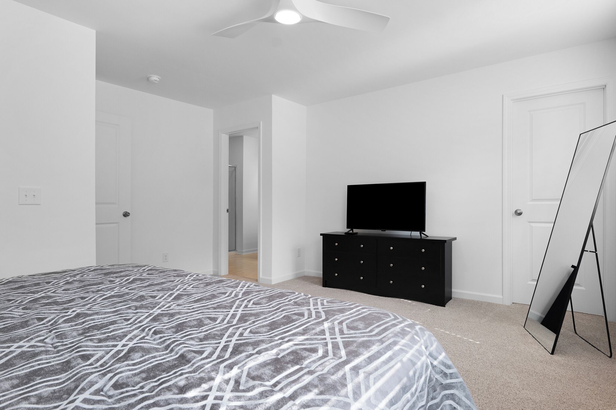 A bedroom with a large bed, a black dresser, a flat-screen TV, and a full-length mirror.