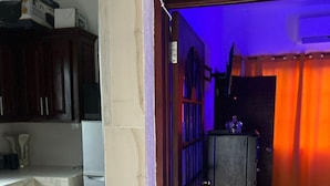 A room with a tiled floor, a door leading to a dark room, and a kitchen area with cabinets and a refrigerator.