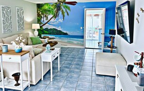 A living room with a beach mural, a sofa, a coffee table, a side table, and a small table with decorative items. - Heart of The Villages, Near Sumter Landing! 2BR/2BA Golf Cart,Guest IDs included (The Villages)