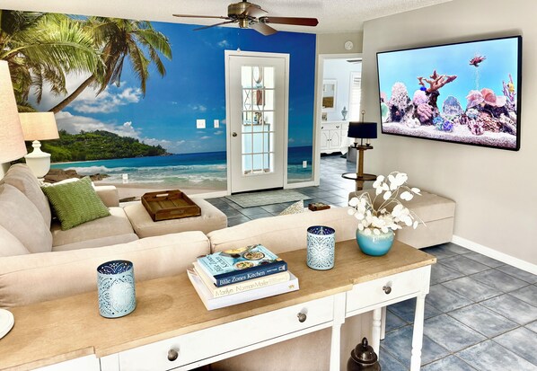A living room with a beach mural, a ceiling fan, a TV, and a coffee table with books and decorative items.
