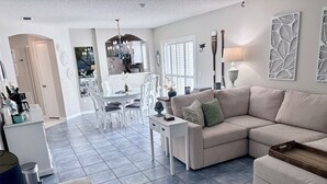 Una sala moderna con un sofá seccional color beige, un comedor con sillas blancas y una cocina al fondo.