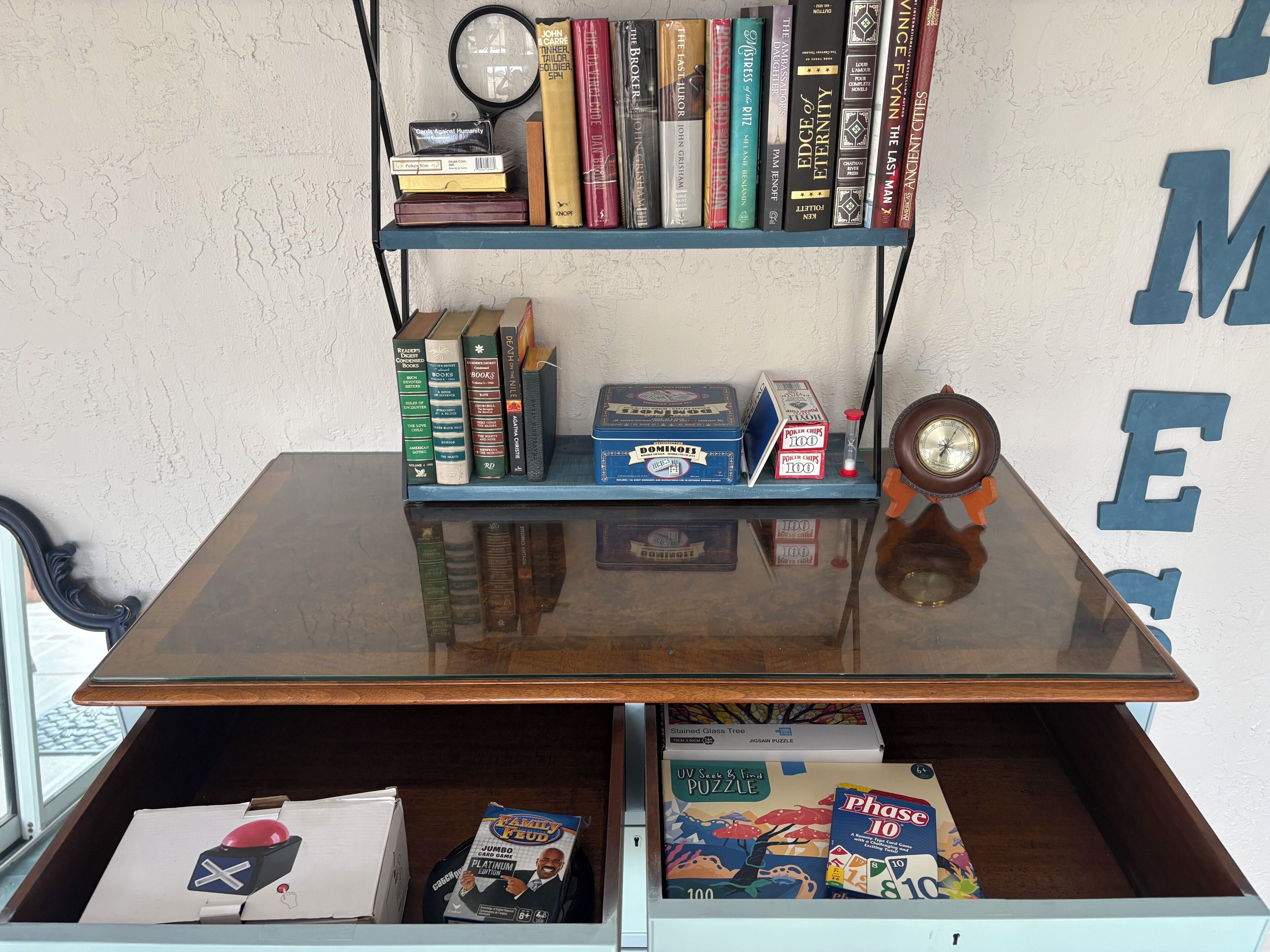 A room with a glass-top table, a bookshelf with books and a magnifying glass, a clock, and a drawer with puzzle boxes.