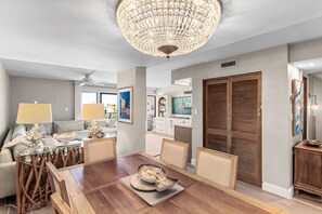A dining area with a wooden table, chairs, a chandelier, and a sofa with cushions. - 2-Story Penthouse - Gulf Views - Private Beach - Crib + Baby Gear - SILVER DUNES (Destin)