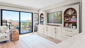 Ein modernes Wohnzimmer mit einem großen Schiebeglasfenster, das auf einen Balkon führt, einem weißen Sofa, einem gemusterten Teppich und einer Kommode mit Dekorationen.