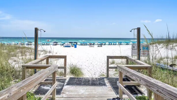 Ein hölzerner Steg führt zu einem Strand mit Sonnenschirmen und Liegestühlen.