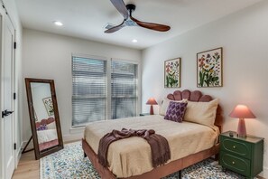 Un dormitorio con cama, dos lámparas de noche, un ventilador de techo y dos ventanas con persianas.