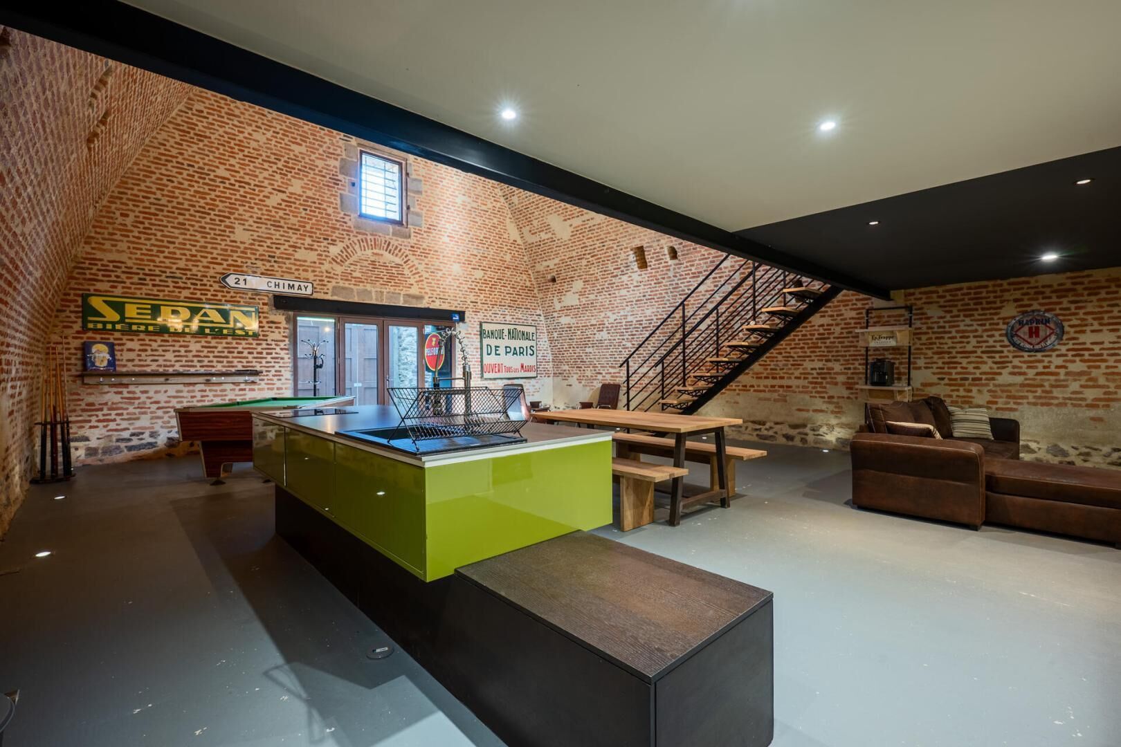 A billiards room with a pool table, a green bar counter, a staircase, and a seating area with sofas.