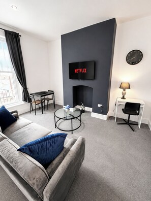 A modern living room with a sofa, a coffee table, a fireplace, a TV mounted on the wall, and a dining area with a table and chairs. - The Quayside Serviced Apartments | The Retreat (Gloucestershire)