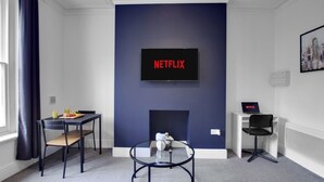 Habitació amb un televisor fixat a la paret que mostra “NETFLIX”, una taula petita amb cadirees, una taula rodona i una llar de foc.