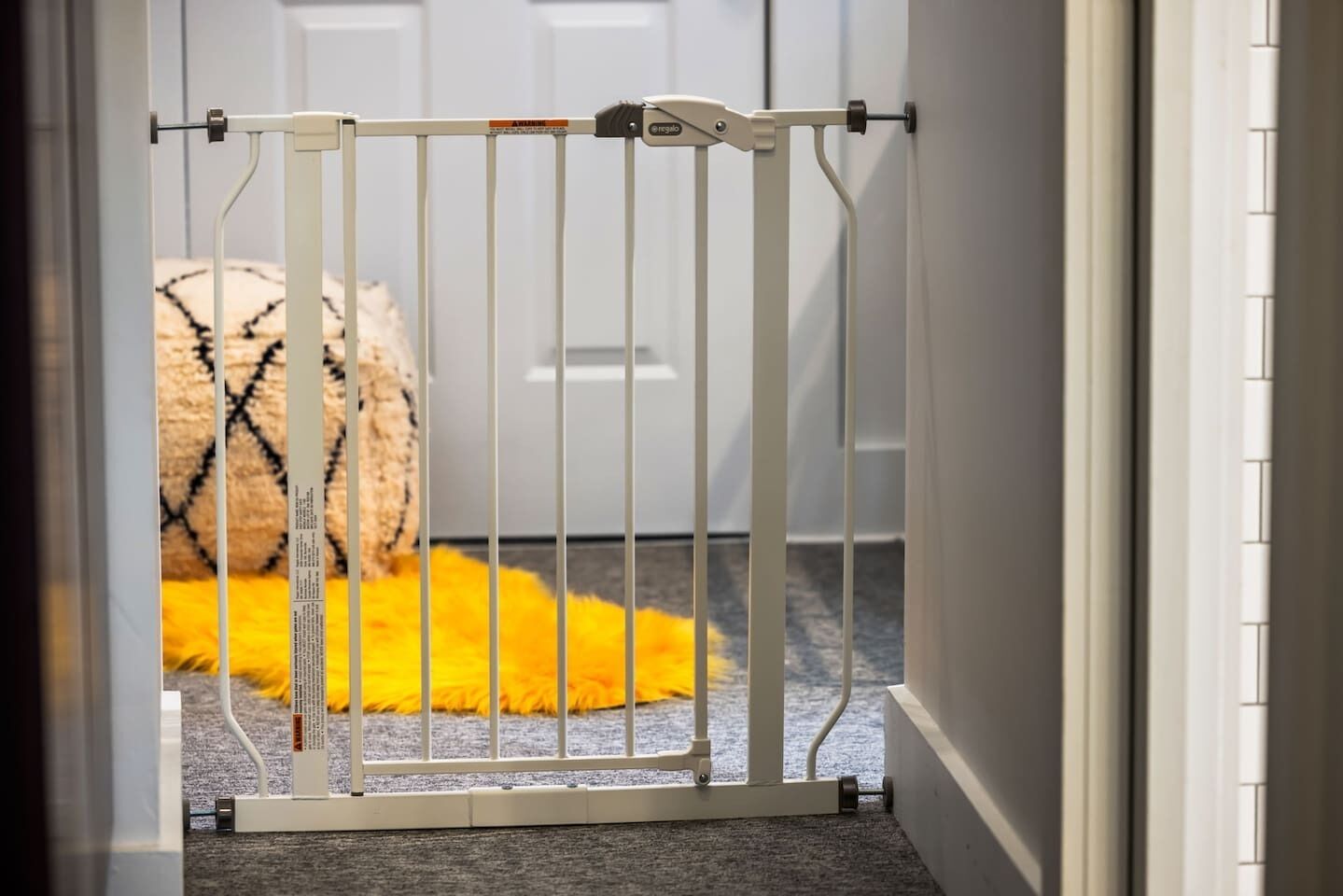 Une barrière de sécurité blanche est installée dans un passage, avec un tapis jaune moelleux et un coussin à motifs en arrière-plan.