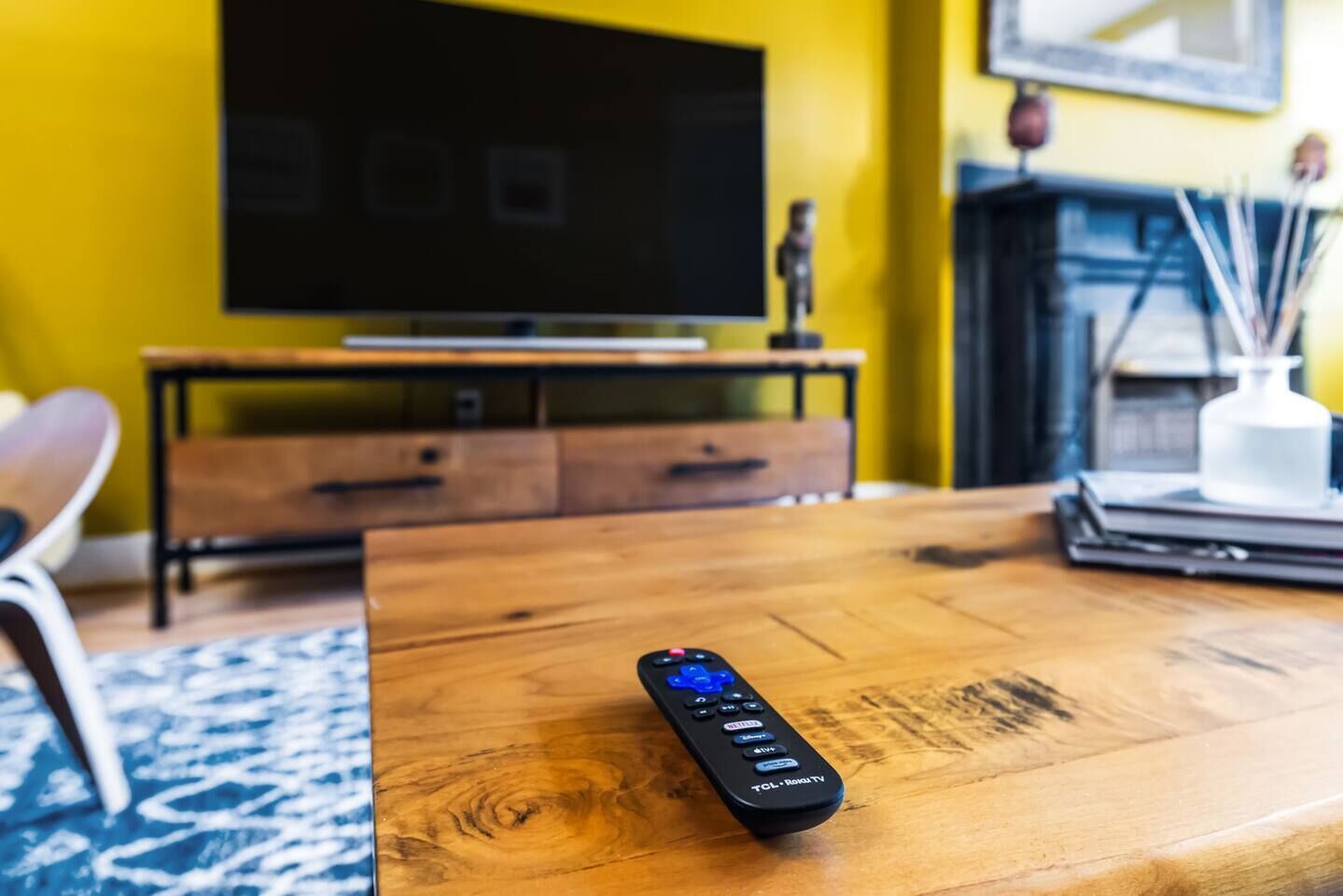 Une télécommande posée sur une table basse en bois dans un salon, avec un téléviseur, une cheminée et un mur jaune.