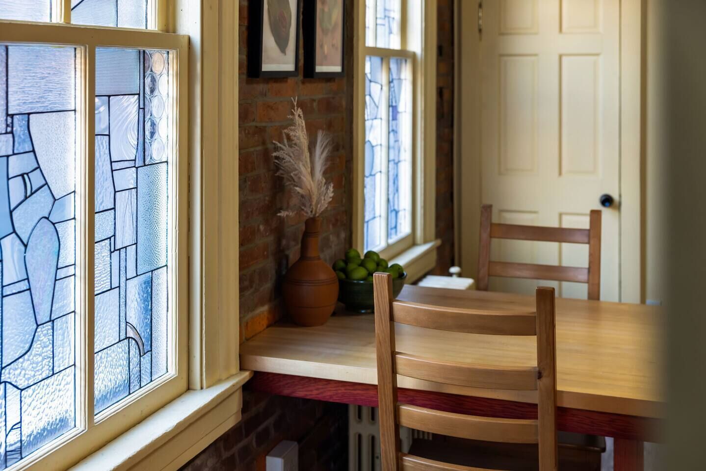 A dining area with a wooden table and chairs, a vase with dried plants, and a brick wall with a window with stained glass.
