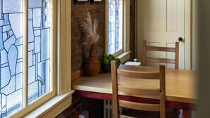 Un coin repas avec une table et des chaises en bois, un vase contenant des plantes séchées et un mur de briques agrémenté d’une fenêtre aux vitraux.