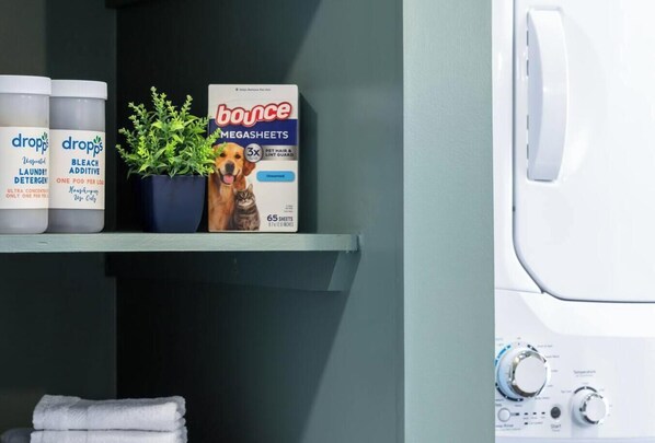 A laundry area with a shelf holding bottles of laundry detergent, a box of Bounce Megasheets, and a potted plant. - Modern Traveler`s Loft| Walkable, Pet-Friendly (Pittsburgh)