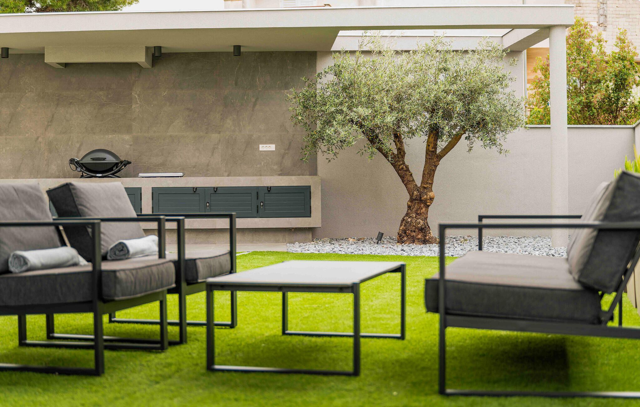 A modern outdoor seating area with a sofa, chairs, and a table, a tree, and a grill against a concrete wall.