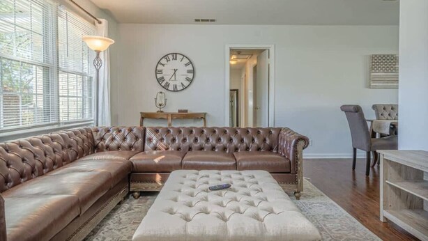 A living room with a brown leather sectional sofa, a white ottoman, a large wall clock, and a dining area with a chair and table. - Charter Oak's Retreat- Near Ft. Benning - Perfect for Graduations/Best Ranger (Columbus)