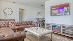 A living room with a brown leather sofa, a white ottoman, a fireplace, and a mounted TV displaying a channel guide. - Charter Oak's Retreat- Near Ft. Benning - Perfect for Graduations/Best Ranger (Columbus)