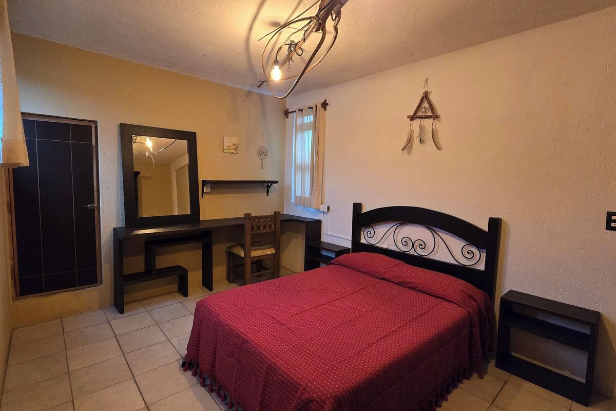 A bedroom with a bed, a desk, a chair, a mirror, a wardrobe, and a hanging light fixture.