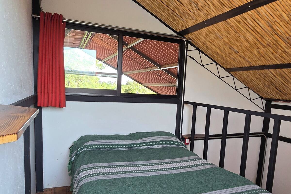 A small, cozy room with a wooden floor, a bed with a green and white striped blanket, a window with red curtains, and a bamboo ceiling.