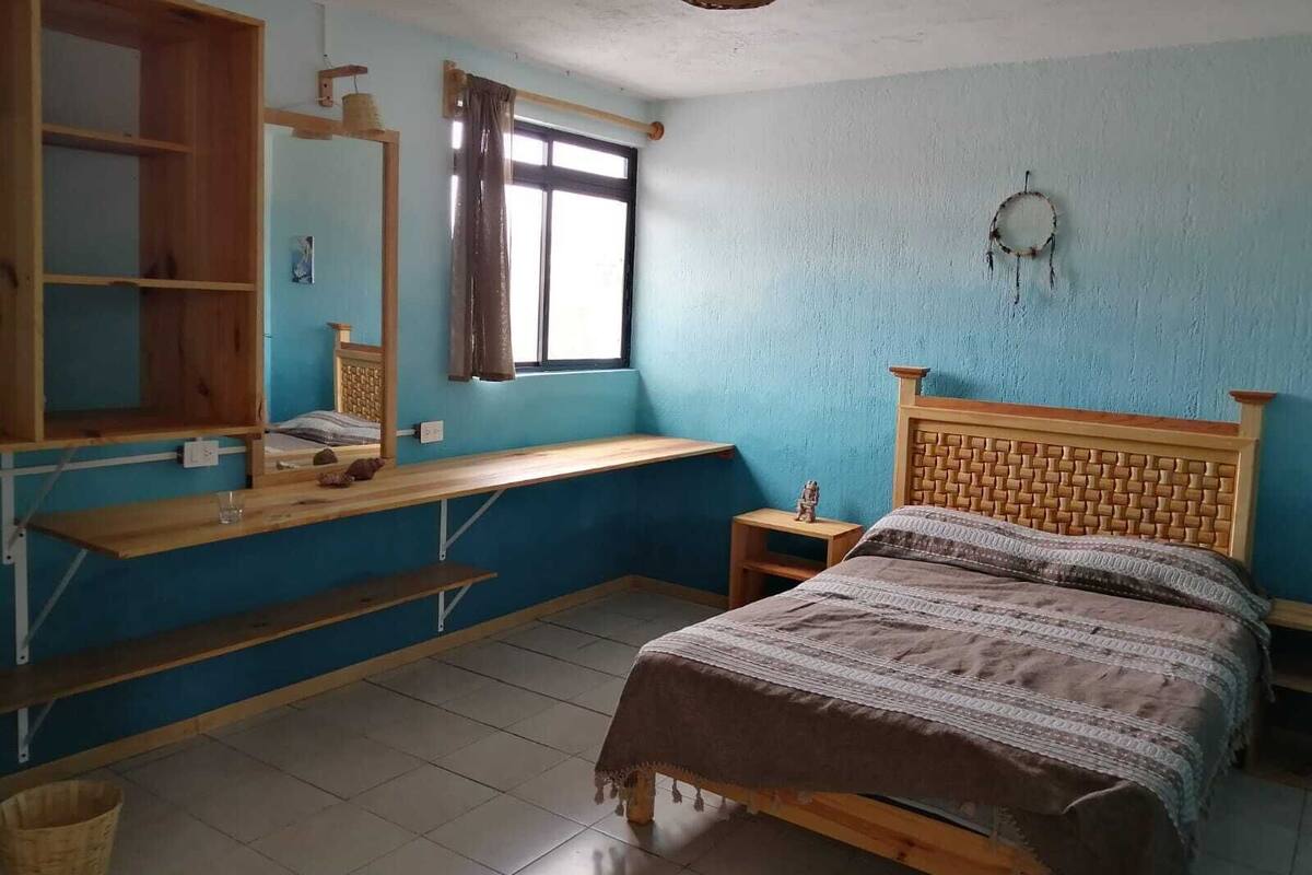 A bedroom with a wooden bed, a wooden desk, a window with a curtain, a wall-mounted dreamcatcher, and a woven basket on the ceiling.