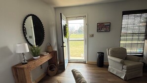 Une pièce comprenant une console en bois, une chaise, un banc, un grand miroir et une fenêtre avec des stores.