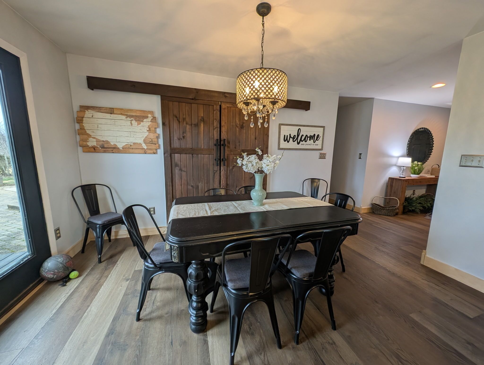 Un comedor con una mesa de madera oscura, sillas negras, una araña y un cartel con la palabra 'welcome'.