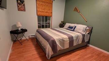 Een slaapkamer met een bed, een plafondventilator, een raam met zonwering, een klein tafeltje met een lamp en een wandplant.