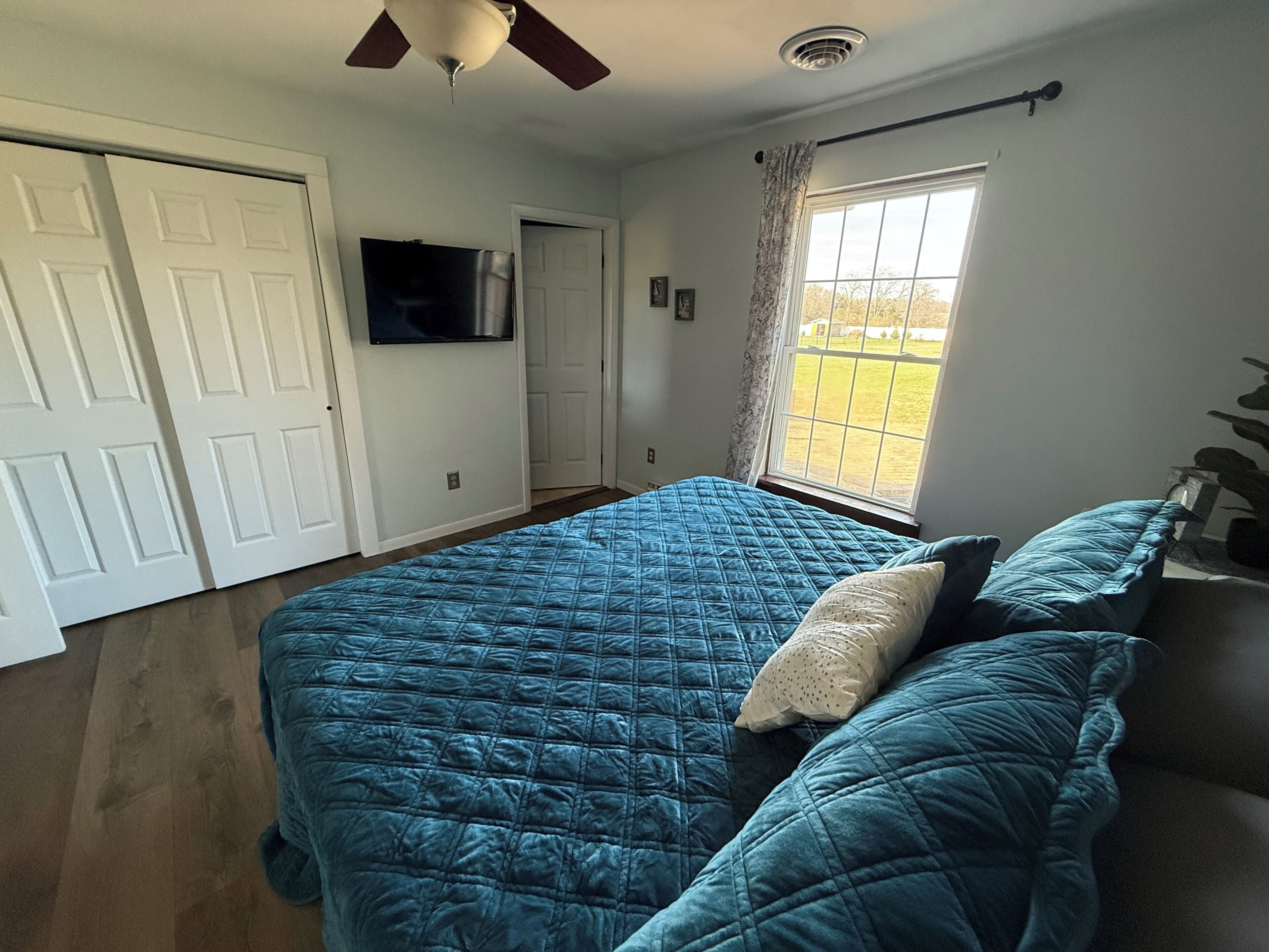 A bedroom with a bed, a ceiling fan, a TV, and a window with a view.