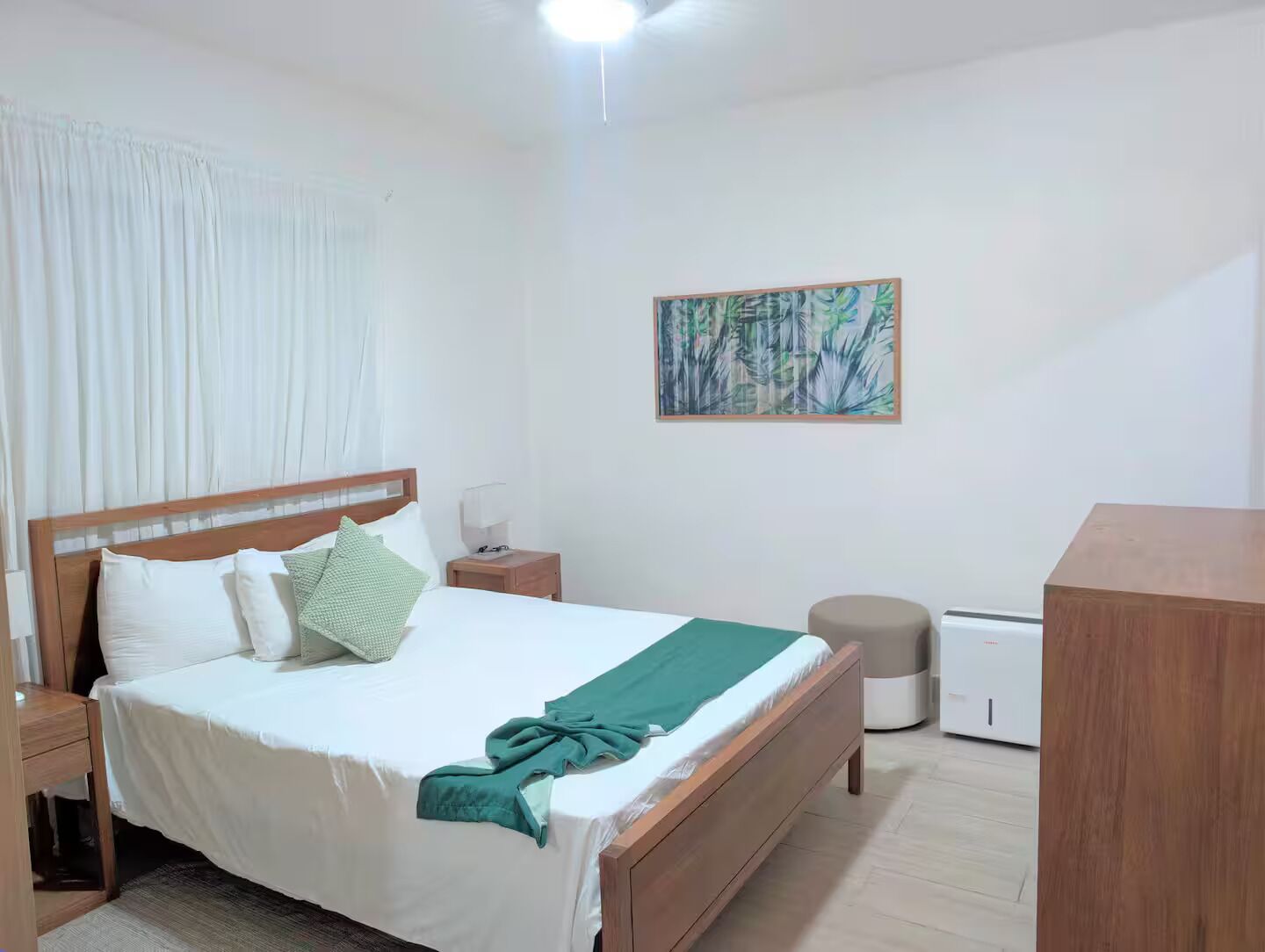 Une chambre avec un lit en bois, du linge de lit blanc, un plaid vert, une table de chevet, une lampe de chevet, un radiateur et un tableau accroché au mur.