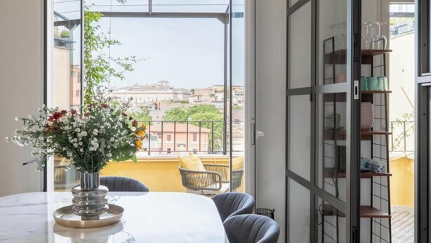 En balkon med udsigt til bygninger, et bord med blomster i en vase og en stol.
